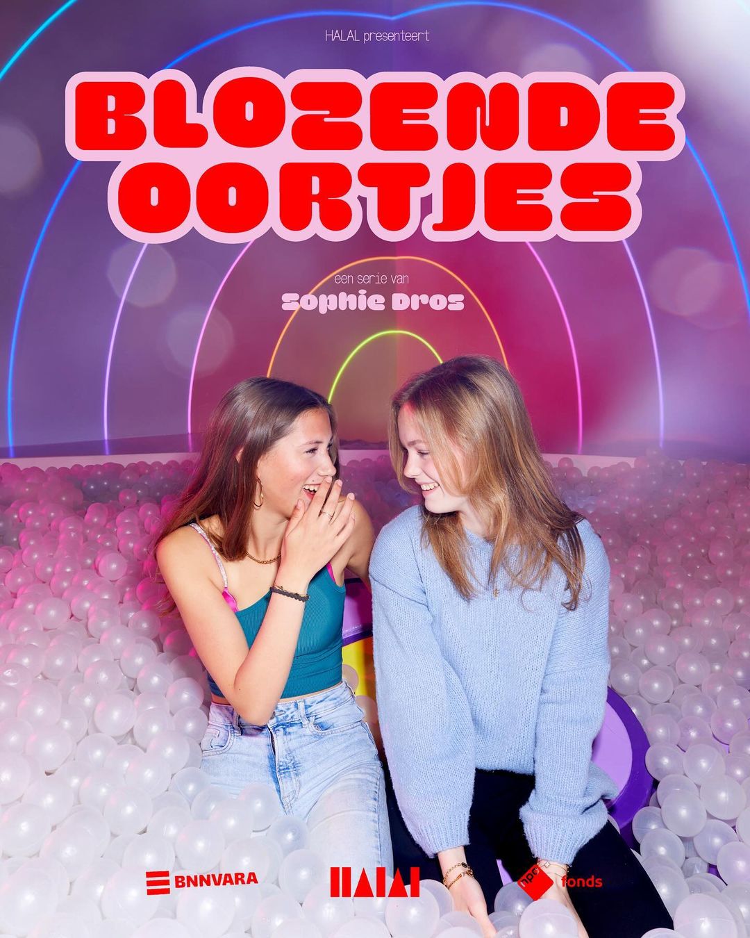 Two young women laugh in a ball pit, promoting a show called 'Blozende Oortjes'. The background features colorful neon archways.