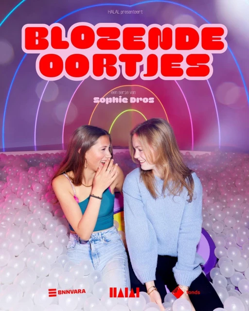 Two young women laugh in a ball pit, promoting a show called 'Blozende Oortjes'. The background features colorful neon archways.