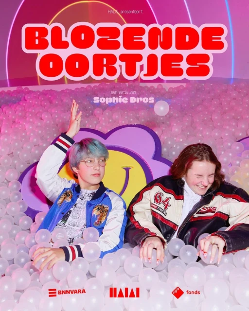 Two young adults in jackets sit in a ball pit with a promotional poster for the show 'Blozende Oortjes' visible behind them.