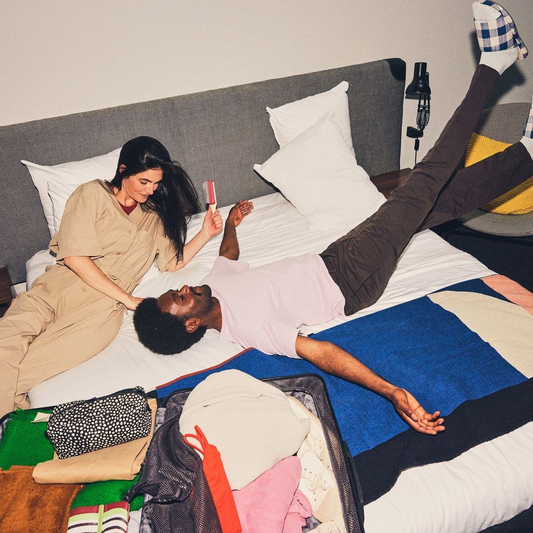 Couple relaxes on a hotel bed, one with legs up in the air and wearing socks, and open suitcase with clothing is in the foreground.