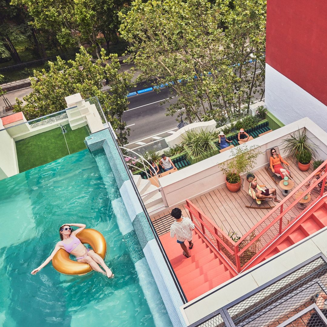 High angle view of a rooftop pool area with a woman floating on an inflatable ring, other people relaxing on a sun deck, and lush trees.