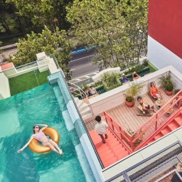 Aerial view of a rooftop terrace with a swimming pool, people lounging, and bright pink stairs by Elza Jo