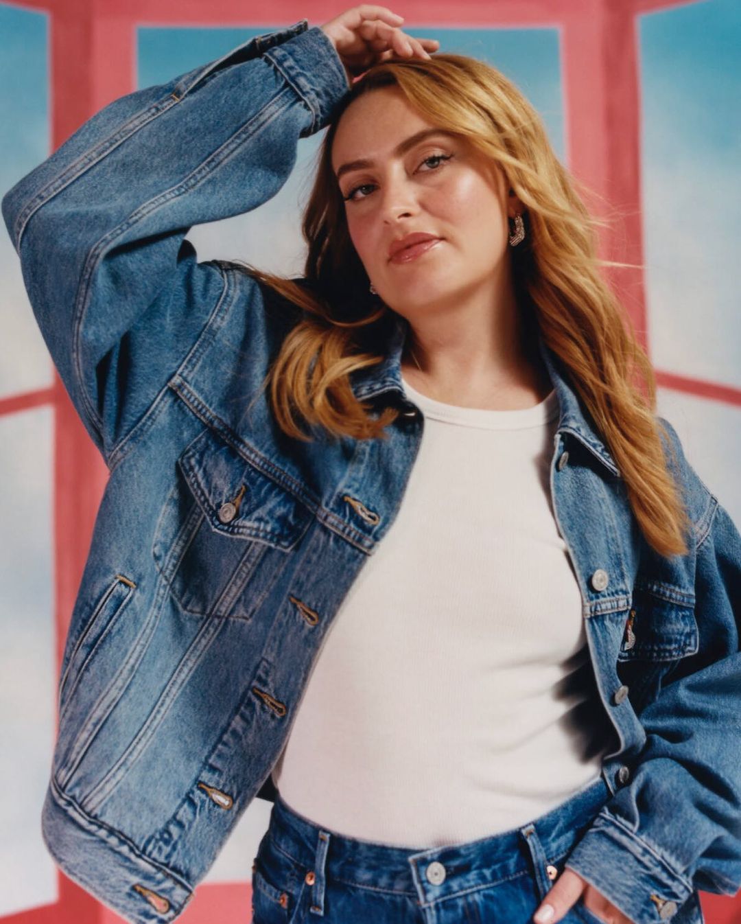 A young woman poses indoors wearing a white t-shirt and full denim outfit, with a denim jacket, jeans, and a pink and blue backdrop.