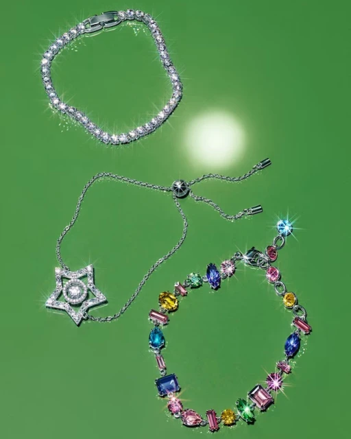 Three silver bracelets with clear and multicolored gemstones are laid out on a green surface, showcasing jewelry design.