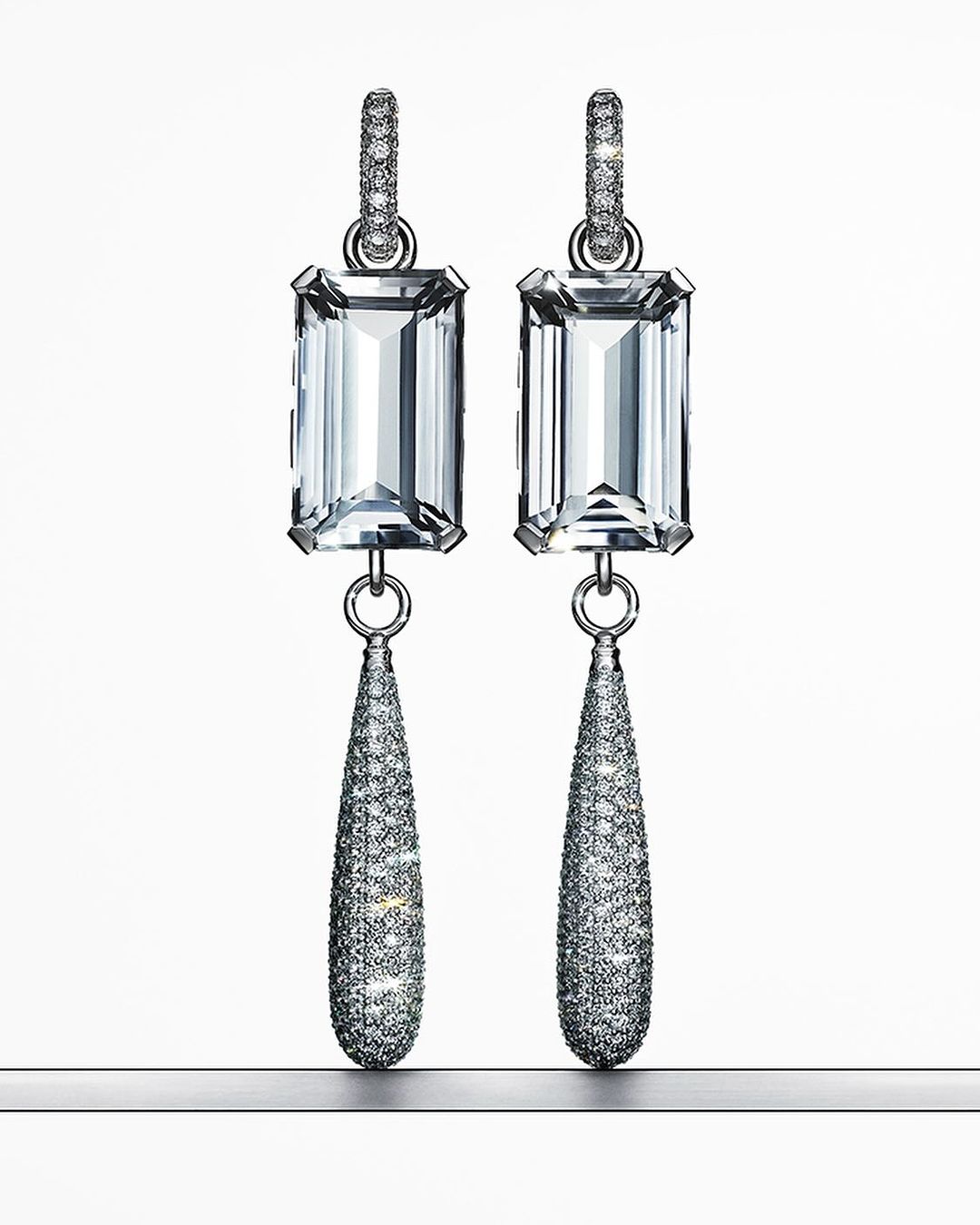 Elegant dangle earrings featuring emerald-cut gemstones and pave diamonds set in silver-tone metal, displayed against a gradient white backdrop.