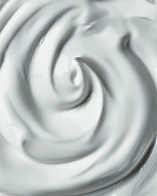 Close-up shot of swirling white cream, lotion, or thick fluid, showcasing its smooth texture and glossy appearance, ideal for beauty or food context.