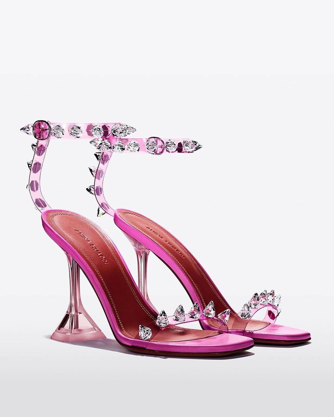 Pair of Amina Muaddi Begum glass-heeled sandals with clear straps, pink leather soles, and jeweled embellishments on a white backdrop.