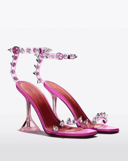 Pair of Amina Muaddi Begum glass-heeled sandals with clear straps, pink leather soles, and jeweled embellishments on a white backdrop.