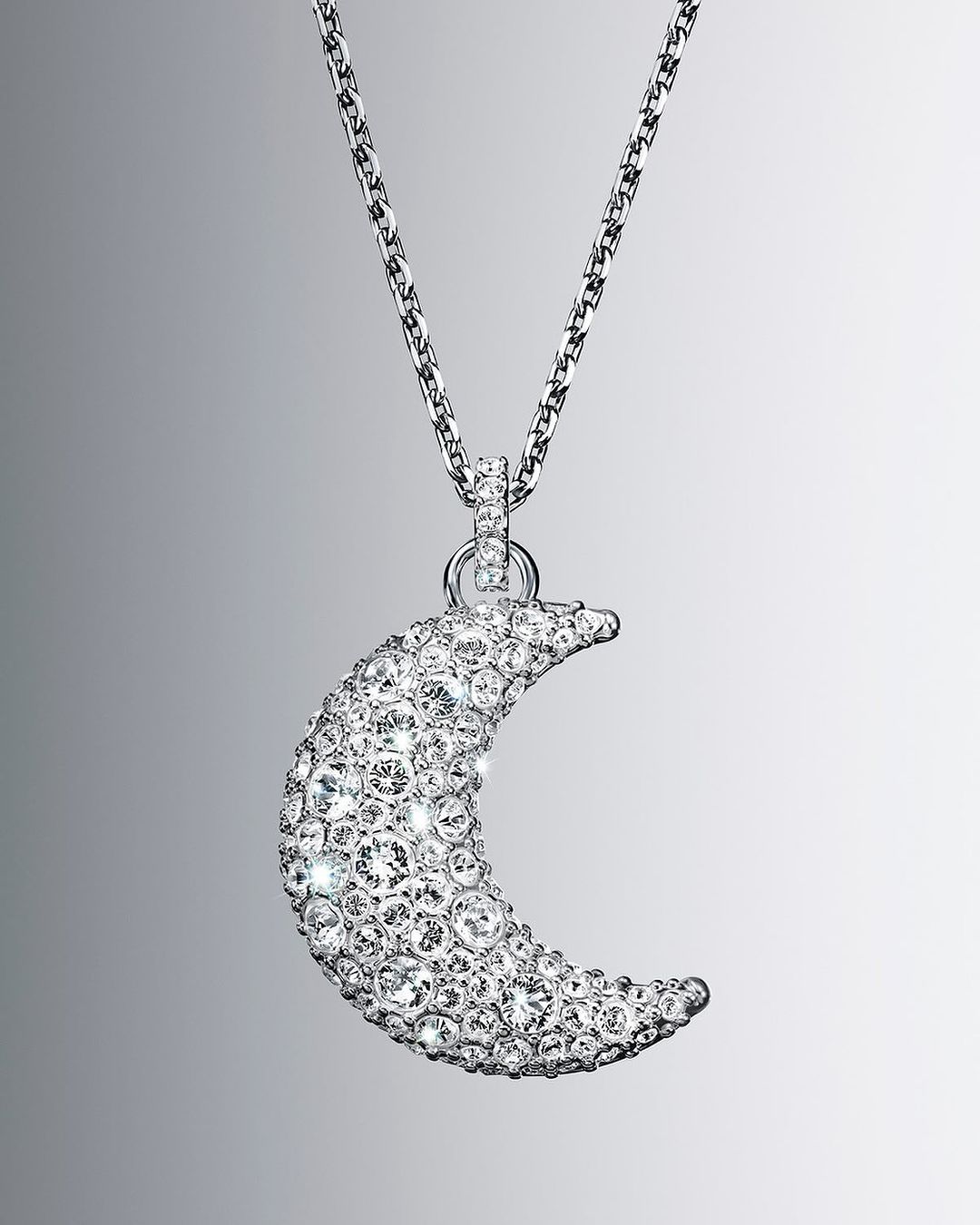 Necklace with a silver chain and a moon-shaped pendant covered in clear crystals, showcased on a neutral grey gradient background.