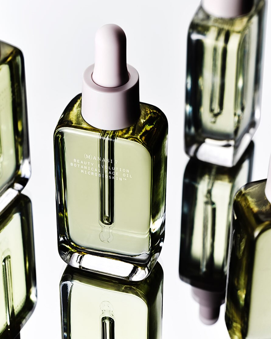 Several square glass bottles of facial oil with pastel pink dropper tops are arranged against a white background and mirrored surface.