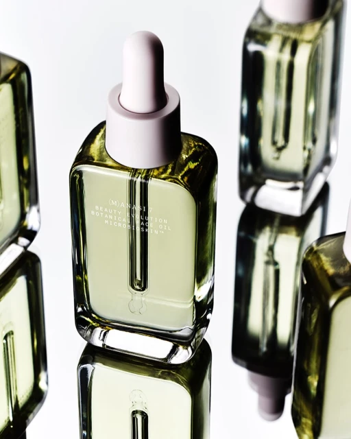 Several square glass bottles of facial oil with pastel pink dropper tops are arranged against a white background and mirrored surface.