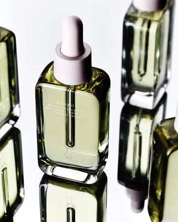 Several square glass bottles of facial oil with pastel pink dropper tops are arranged against a white background and mirrored surface.