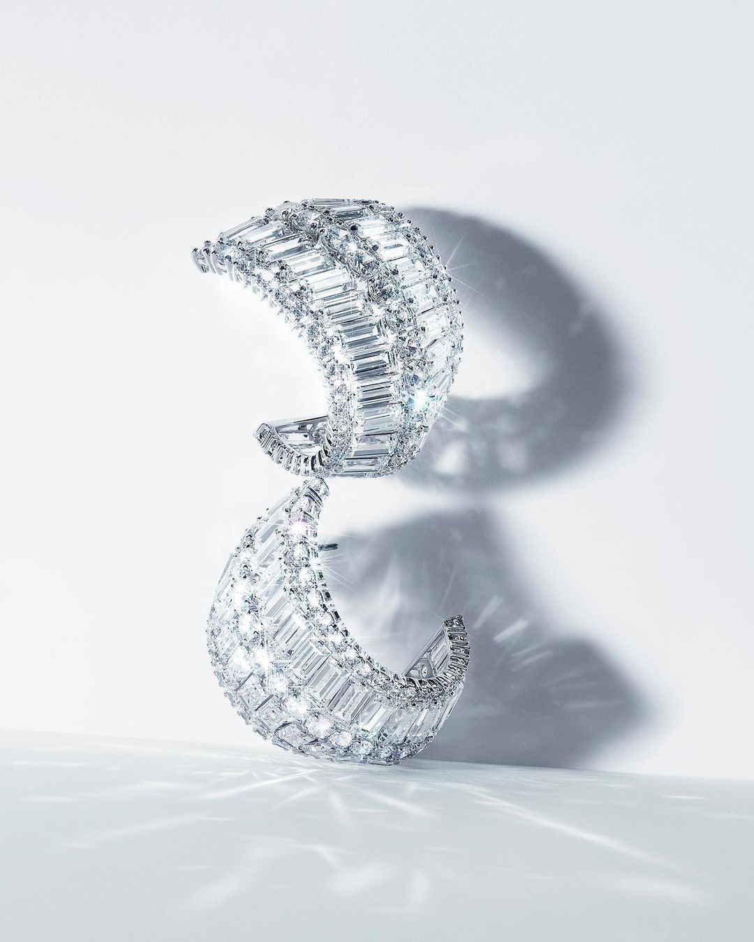 Two crescent-shaped diamond earrings stacked on a white surface with subtle shadows, showcasing their intricate design and sparkle.