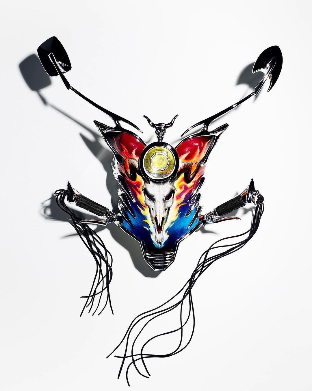 Artistic close-up of a stylized motorcycle fragment with flame-like details and bull skull emblem, shot against a clean white backdrop.