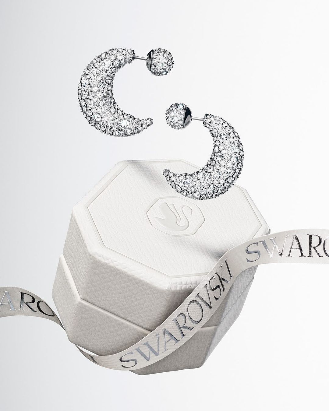 Elevated view of crystal-studded half-hoop earrings displayed over a white gift box, accented by a branded ribbon.