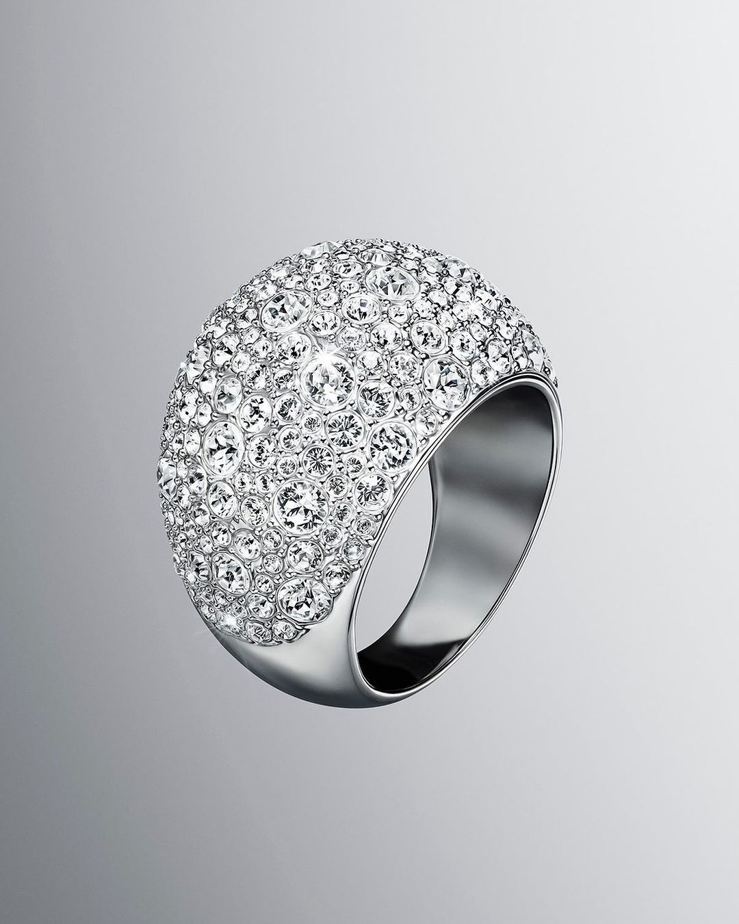 Close-up of a large, dome-shaped ring encrusted with clear crystals, set on a silver metal band against a gradient grey background.