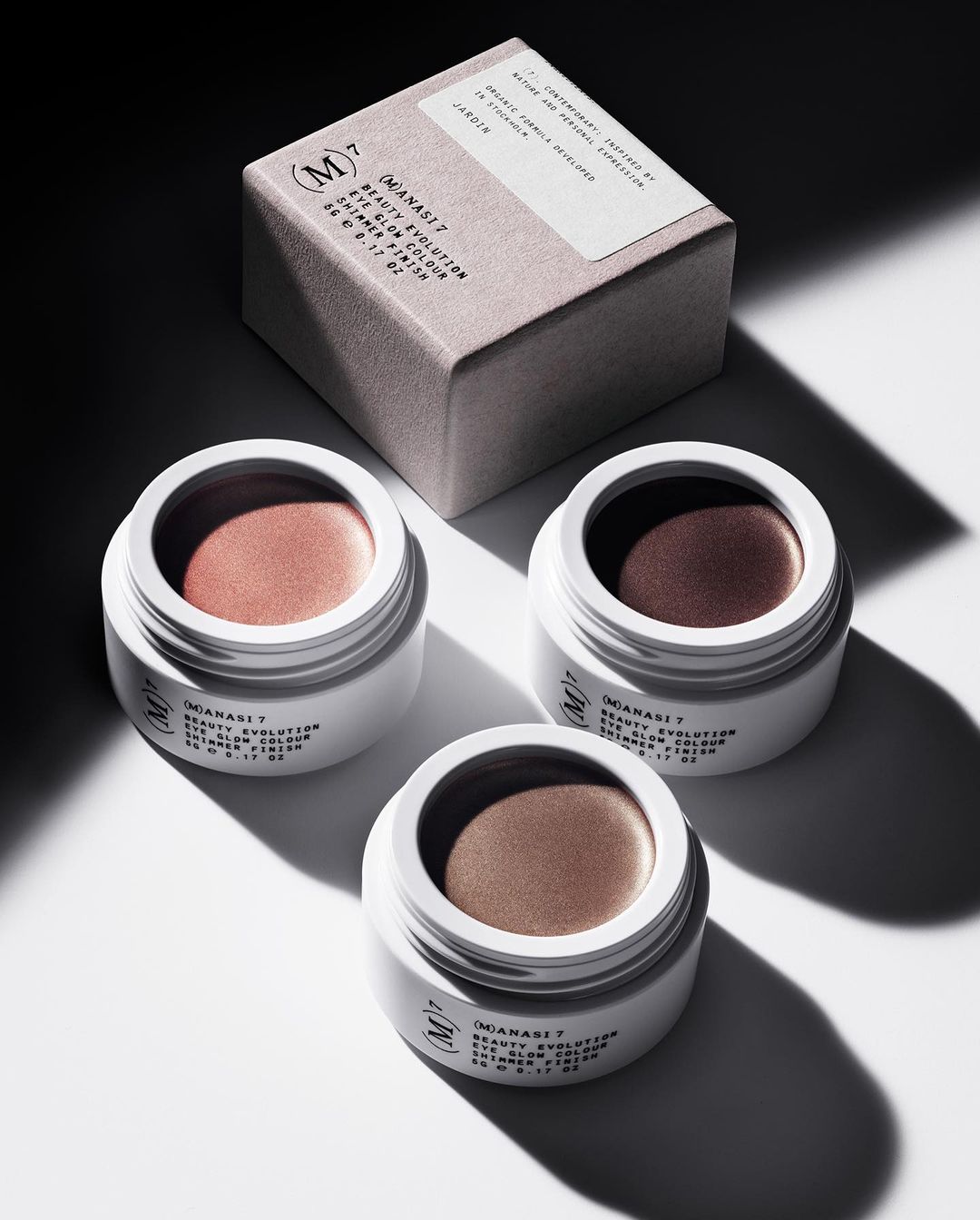 Three cream eyeshadow pots in white containers with product packaging on a white surface in a beauty product studio shot.