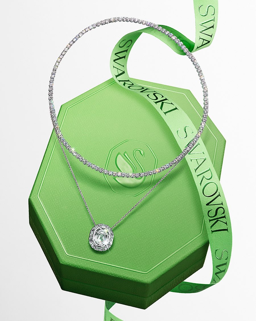 Elegant jewelry display featuring a crystal necklace and pendant on a green Swarovski gift box with a logo ribbon, studio setting.