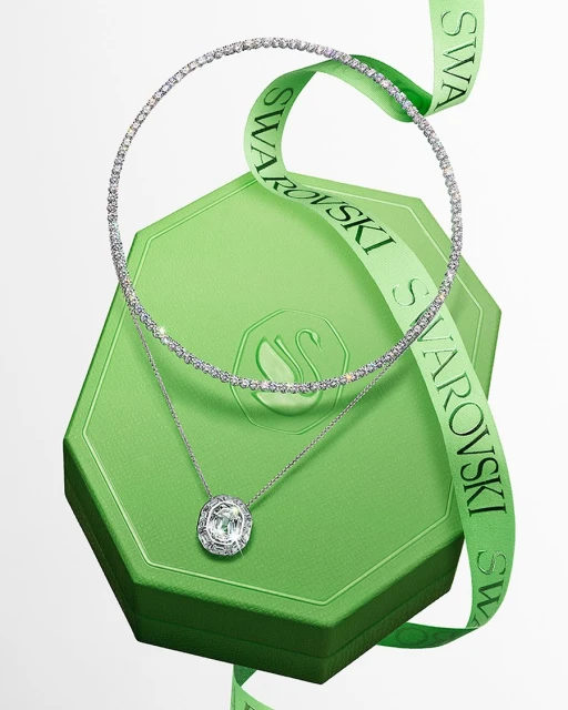 Elegant jewelry display featuring a crystal necklace and pendant on a green Swarovski gift box with a logo ribbon, studio setting.