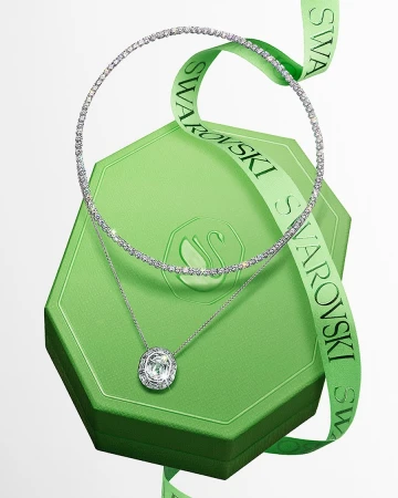 Elegant jewelry display featuring a crystal necklace and pendant on a green Swarovski gift box with a logo ribbon, studio setting.