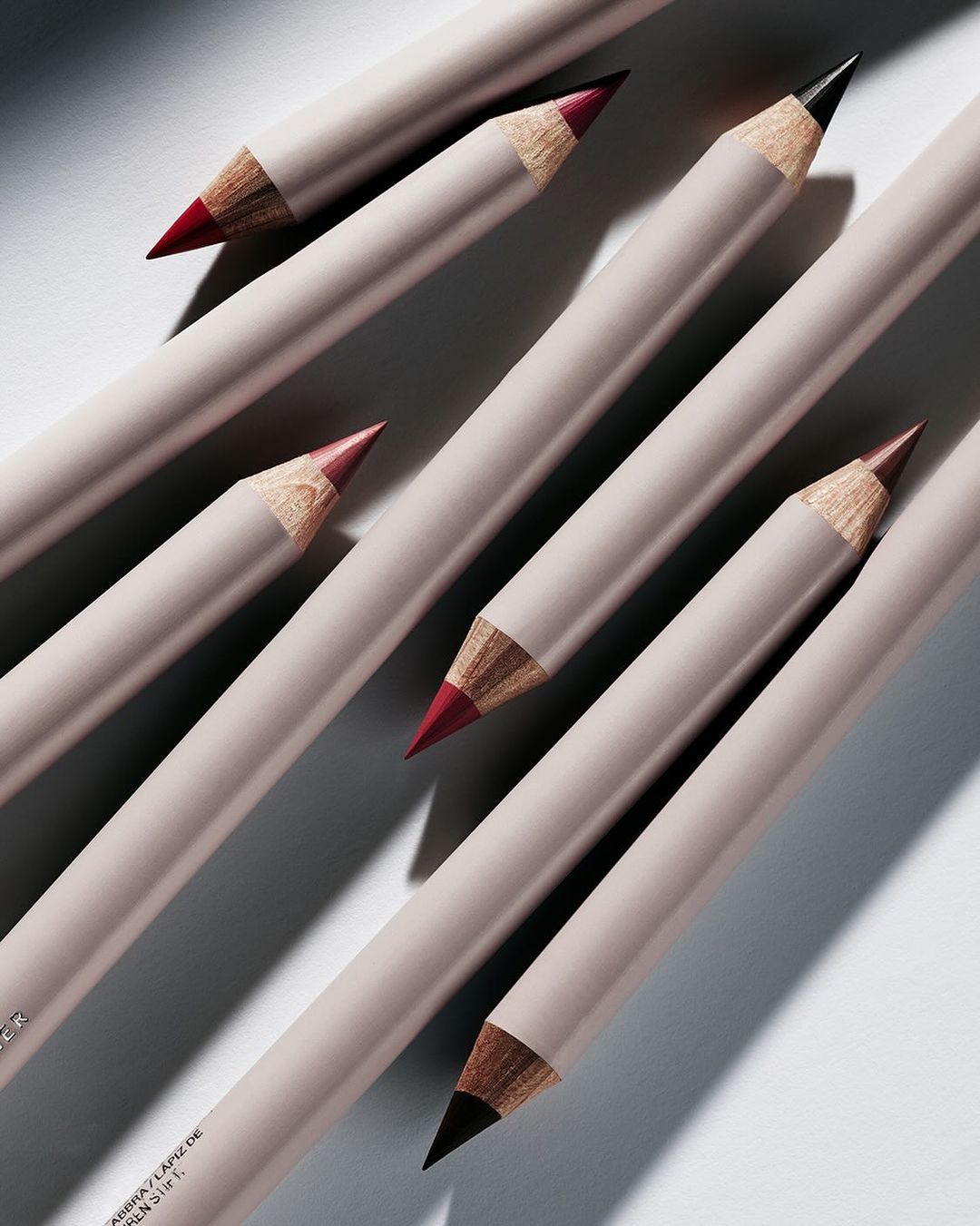 Close-up shot of an arrangement of light-beige colored lip pencils with red and black tips placed on a light-grey surface.