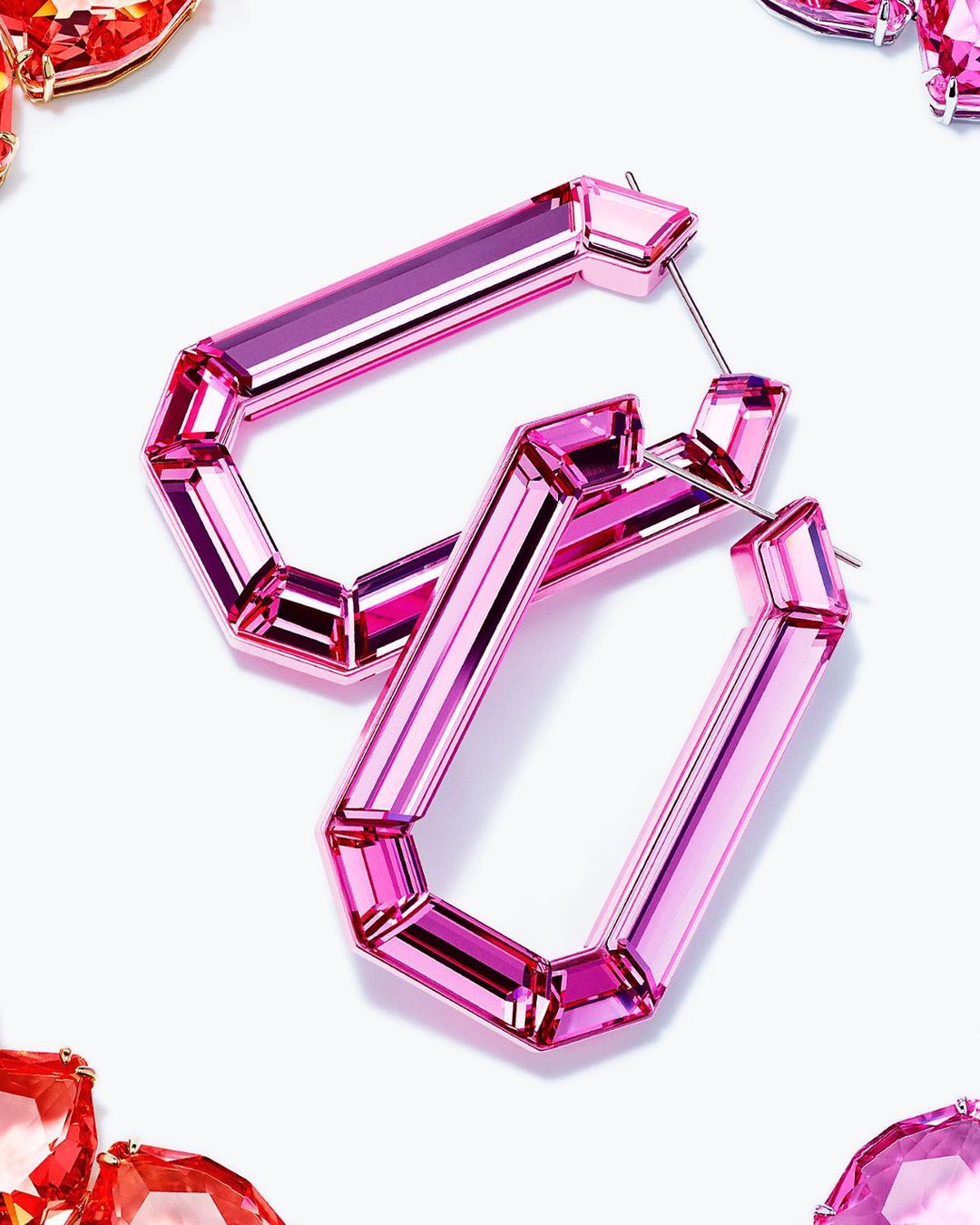 Pink rectangular geometric earrings made of reflective, transparent material laying on white surface with red and pink stones around.