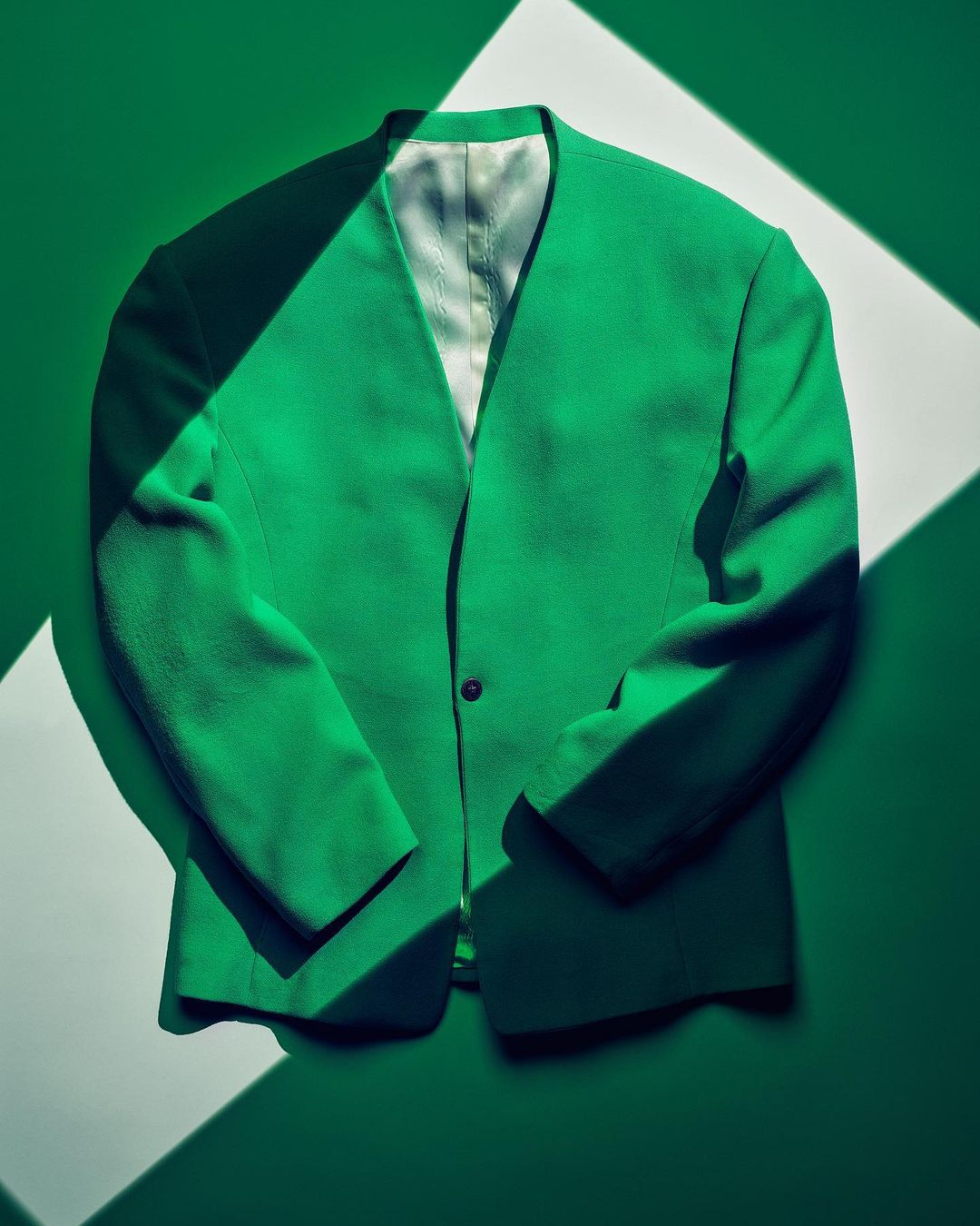 Bright green blazer with a single button closure, displayed against a green background with geometric white patterns created by light.