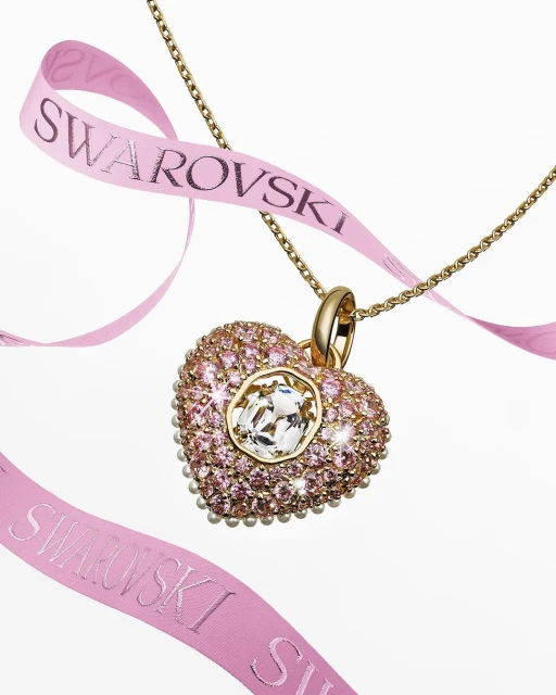 Close-up of a Swarovski crystal heart pendant necklace, with pink ribbon bearing the Swarovski logo, against a white backdrop.