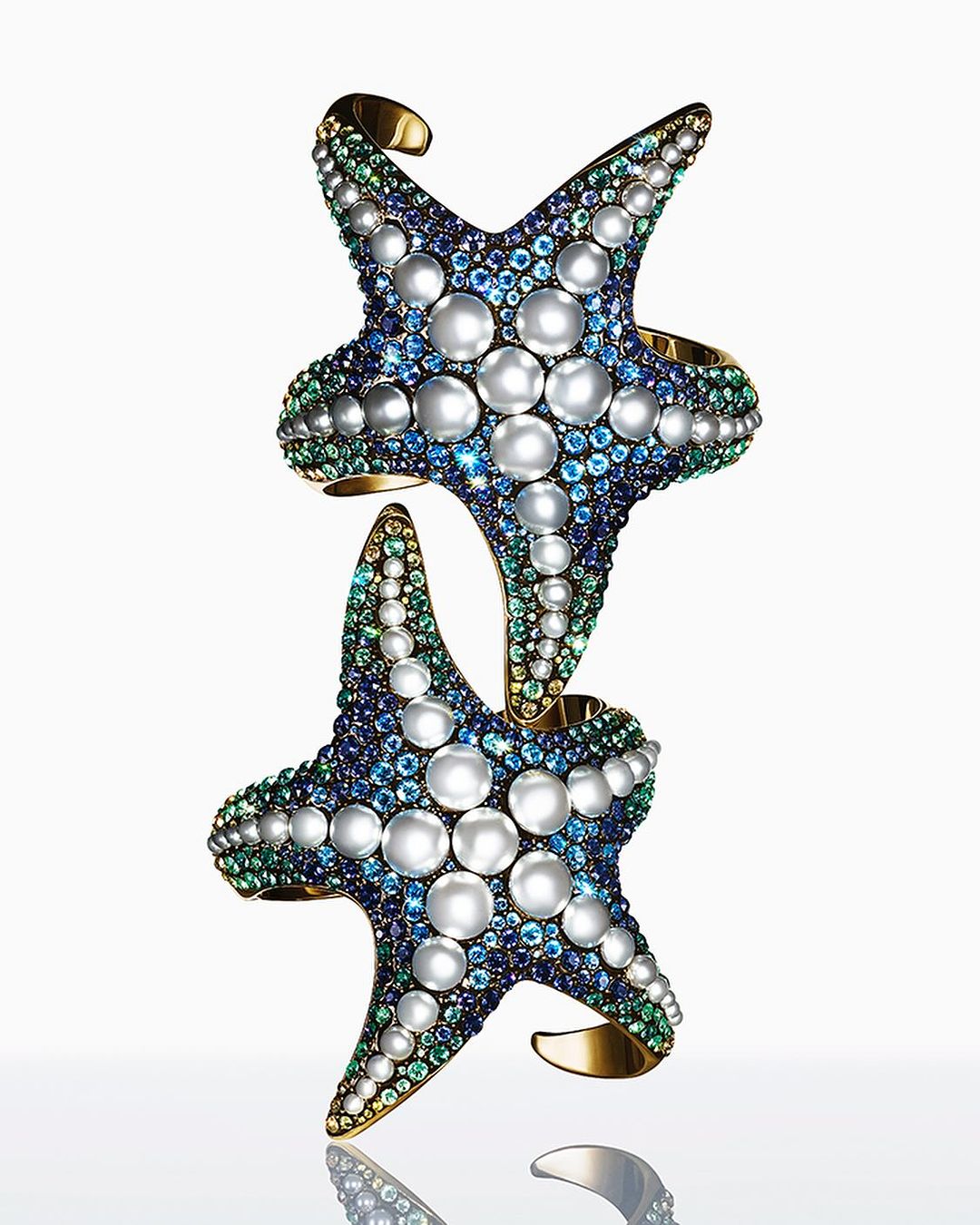 Two starfish cuff bracelets featuring pearls and gemstones, stacked on a reflective surface against a white background. Jewellery still life.
