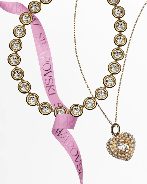 Two Swarovski necklaces, one with crystal bezels and a pink logo ribbon, the other a heart pendant, against a white backdrop.