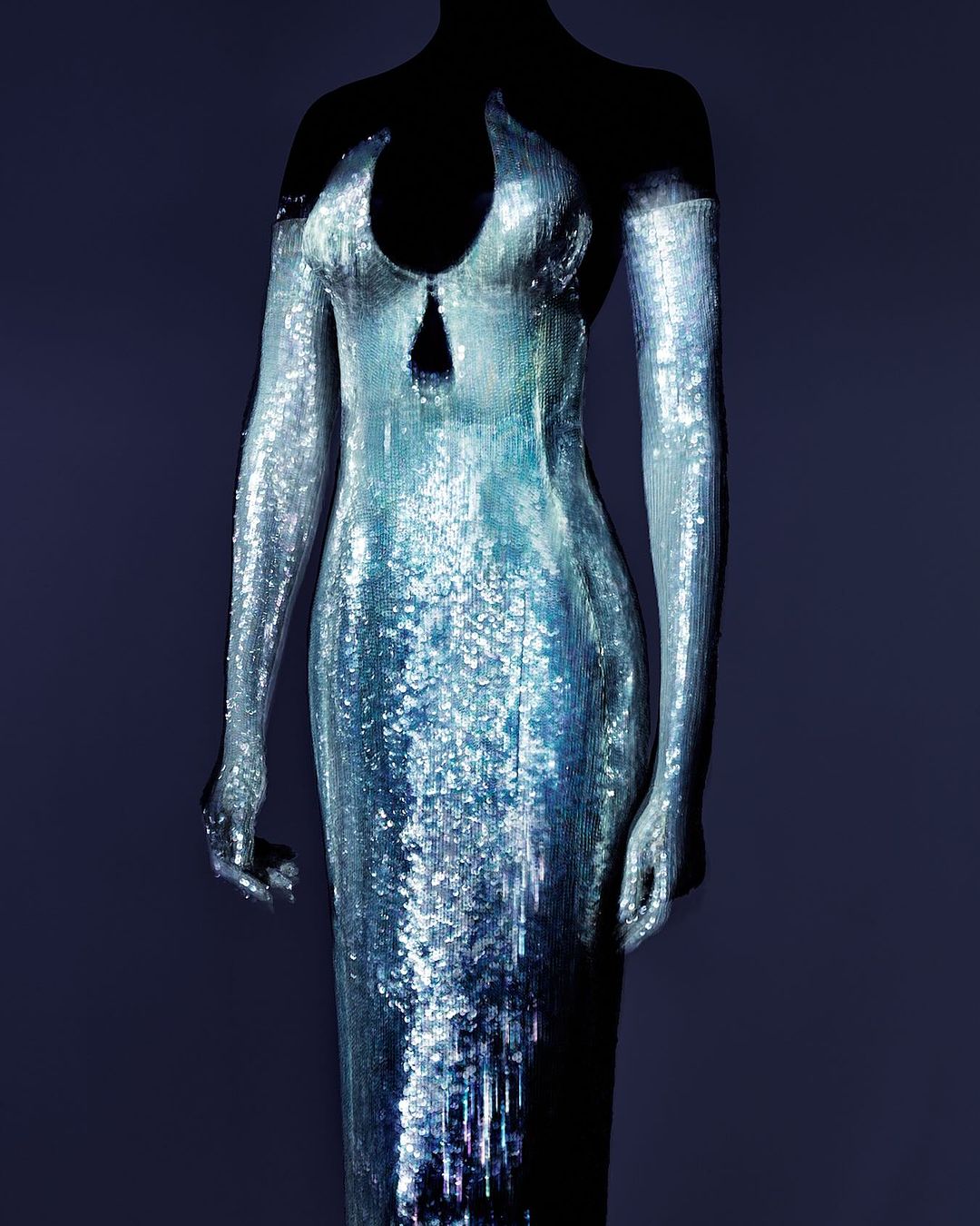 Sequin-covered formal gown on a mannequin against a dark background, silver and blue iridescent fabric with a fitted silhouette.