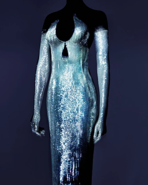 Sequin-covered formal gown on a mannequin against a dark background, silver and blue iridescent fabric with a fitted silhouette.
