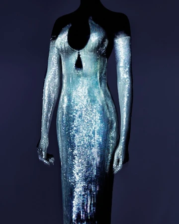 Sequin-covered formal gown on a mannequin against a dark background, silver and blue iridescent fabric with a fitted silhouette.