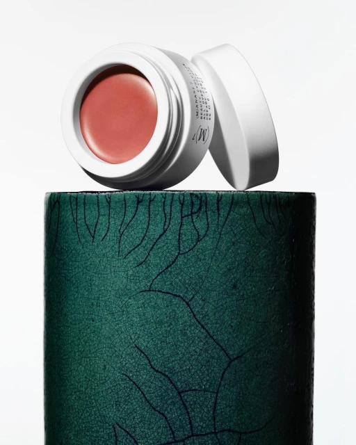 Blush cream in a white container with the lid off and set on a textured green cylinder against a bright white background.