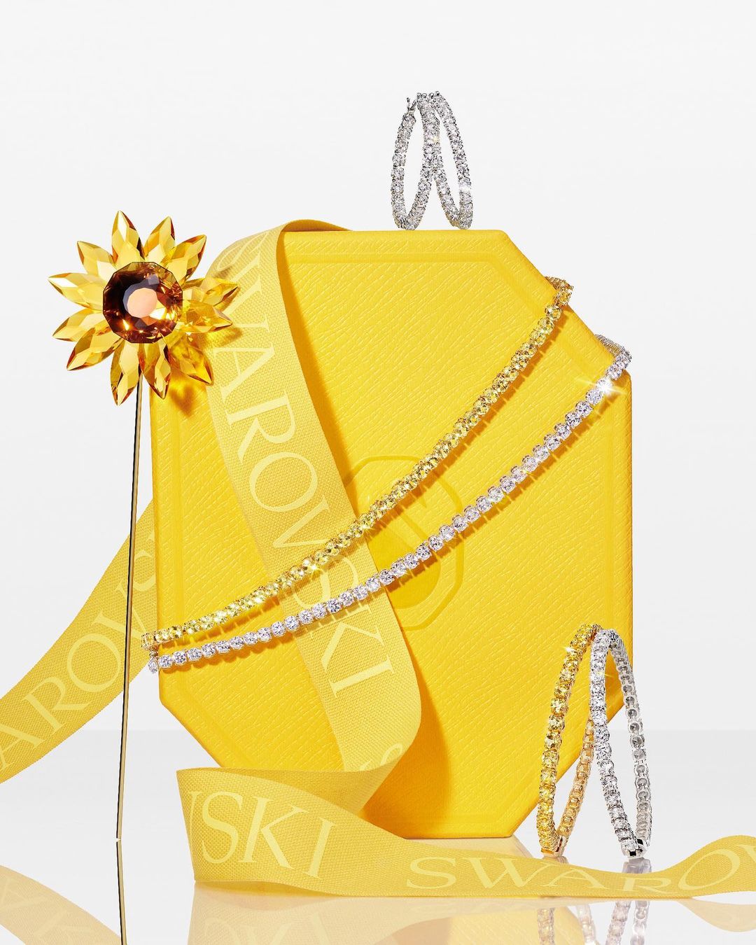 Still life featuring Swarovski jewelry and a yellow box with ribbon, showcasing earrings and necklaces against a bright background.