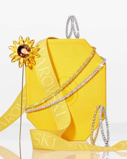 Still life featuring Swarovski jewelry and a yellow box with ribbon, showcasing earrings and necklaces against a bright background.