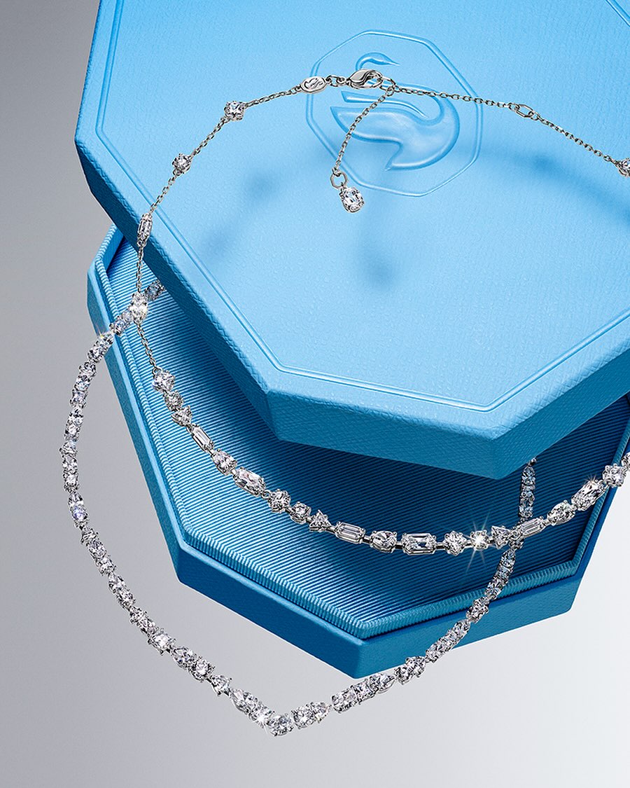 Sparkling crystal necklaces are displayed on a light blue jewelry box against a gradient gray background in this jewelry advertisement.