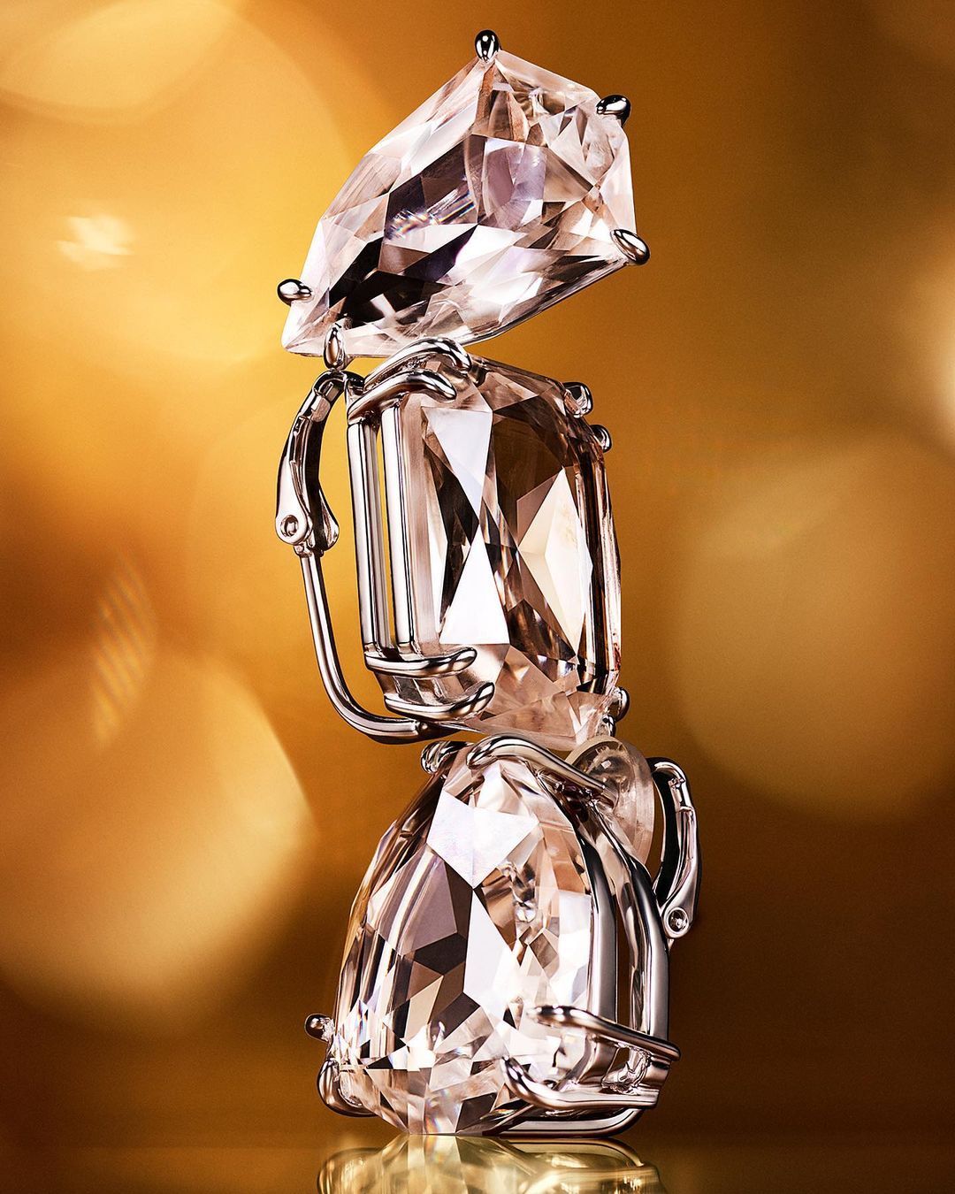 Elegant earring featuring a stack of three differently cut clear gemstones with silver metal hardware against a warm golden backdrop.
