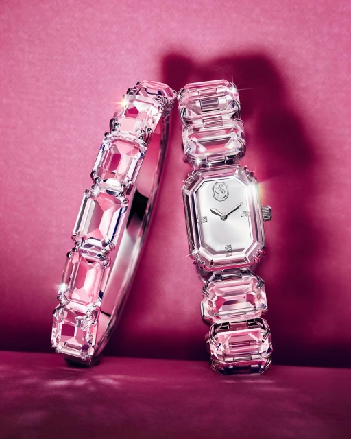 Still life studio shot of a silver-tone metal bracelet and wristwatch encrusted with pink crystal stones against a pink backdrop.
