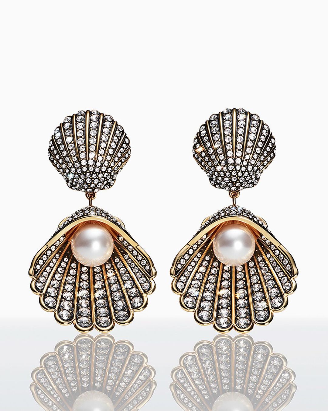 Pair of seashell-shaped dangle earrings with pearl accent and diamond embellishments, studio shot on a bright white reflective surface.