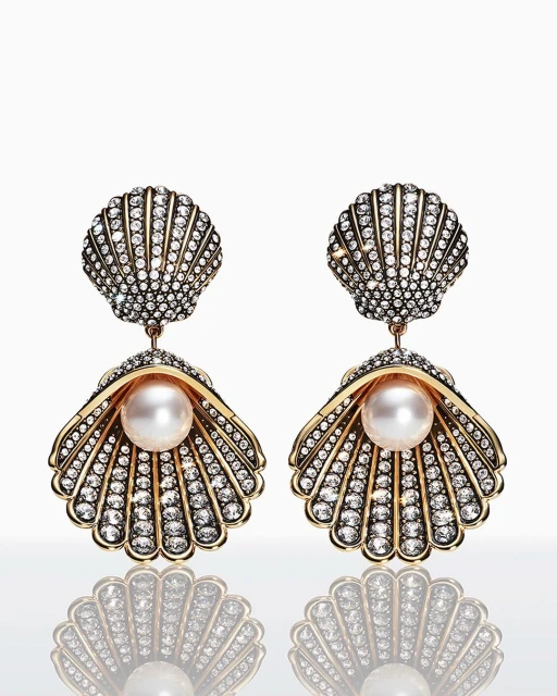 Pair of seashell-shaped dangle earrings with pearl accent and diamond embellishments, studio shot on a bright white reflective surface.