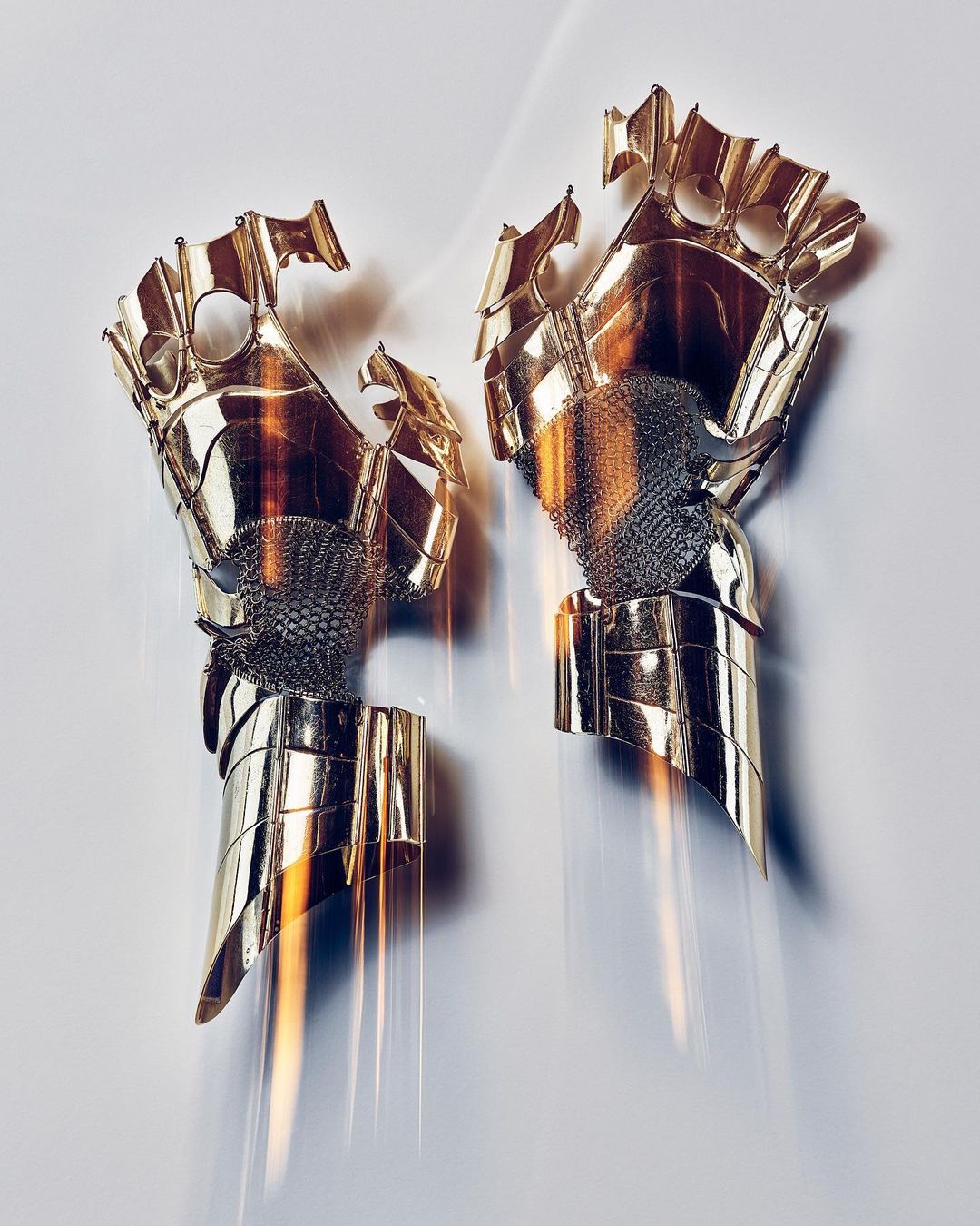 Two mirrored metallic gauntlets with gold and silver coloring displayed against a neutral background, reflecting light and shadow.