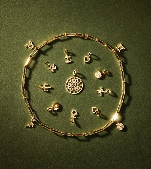 Close-up of a golden chain with various zodiac sign charms scattered on a dark green background, creating a fashionable jewelry display.