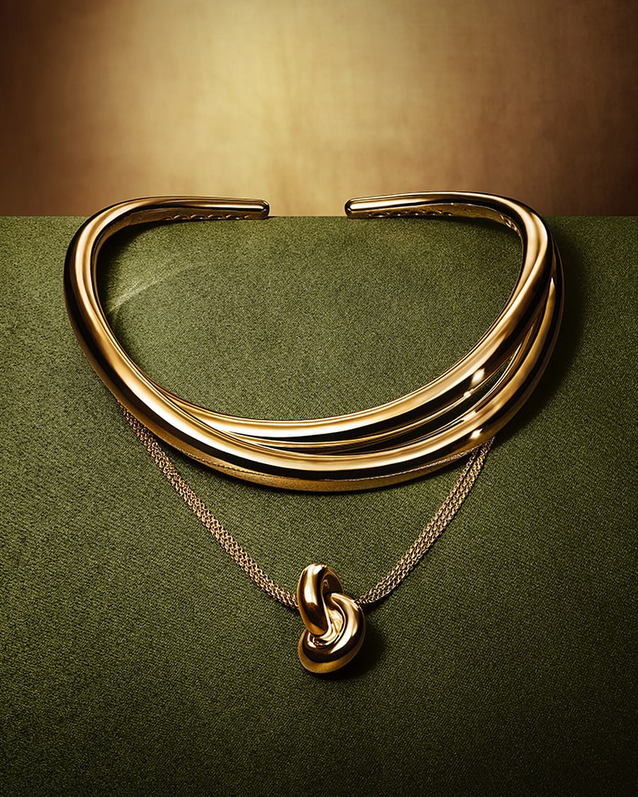 A gold knotted pendant necklace and matching cuff sit on a green surface against a graduated brown background.