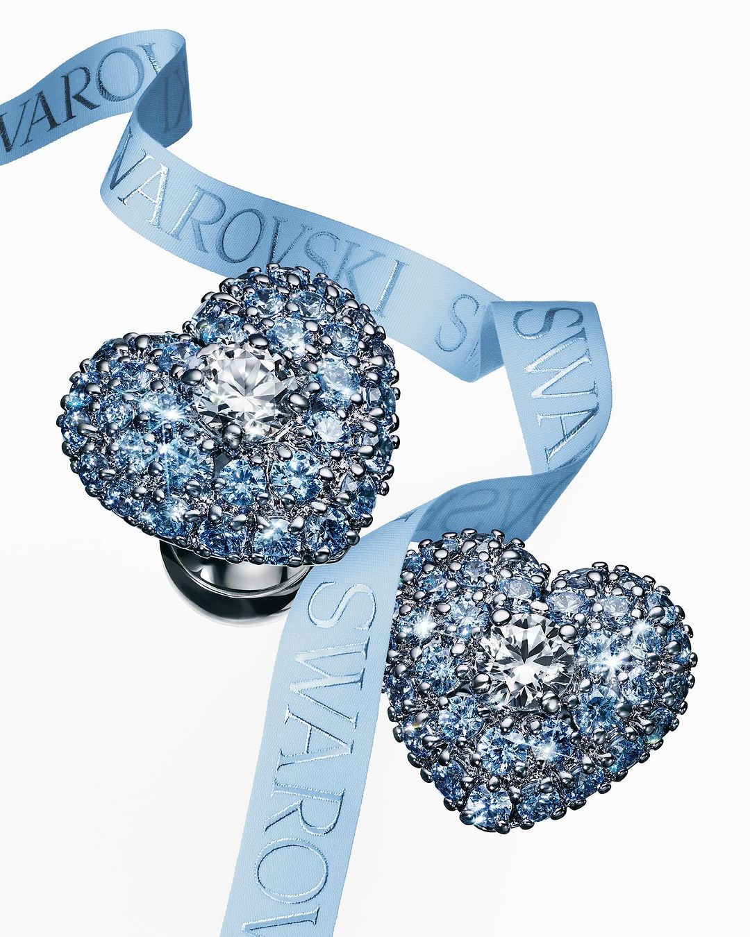 Two heart-shaped Swarovski crystal earrings on a light blue ribbon with the brand name, showcased on a clean white background.