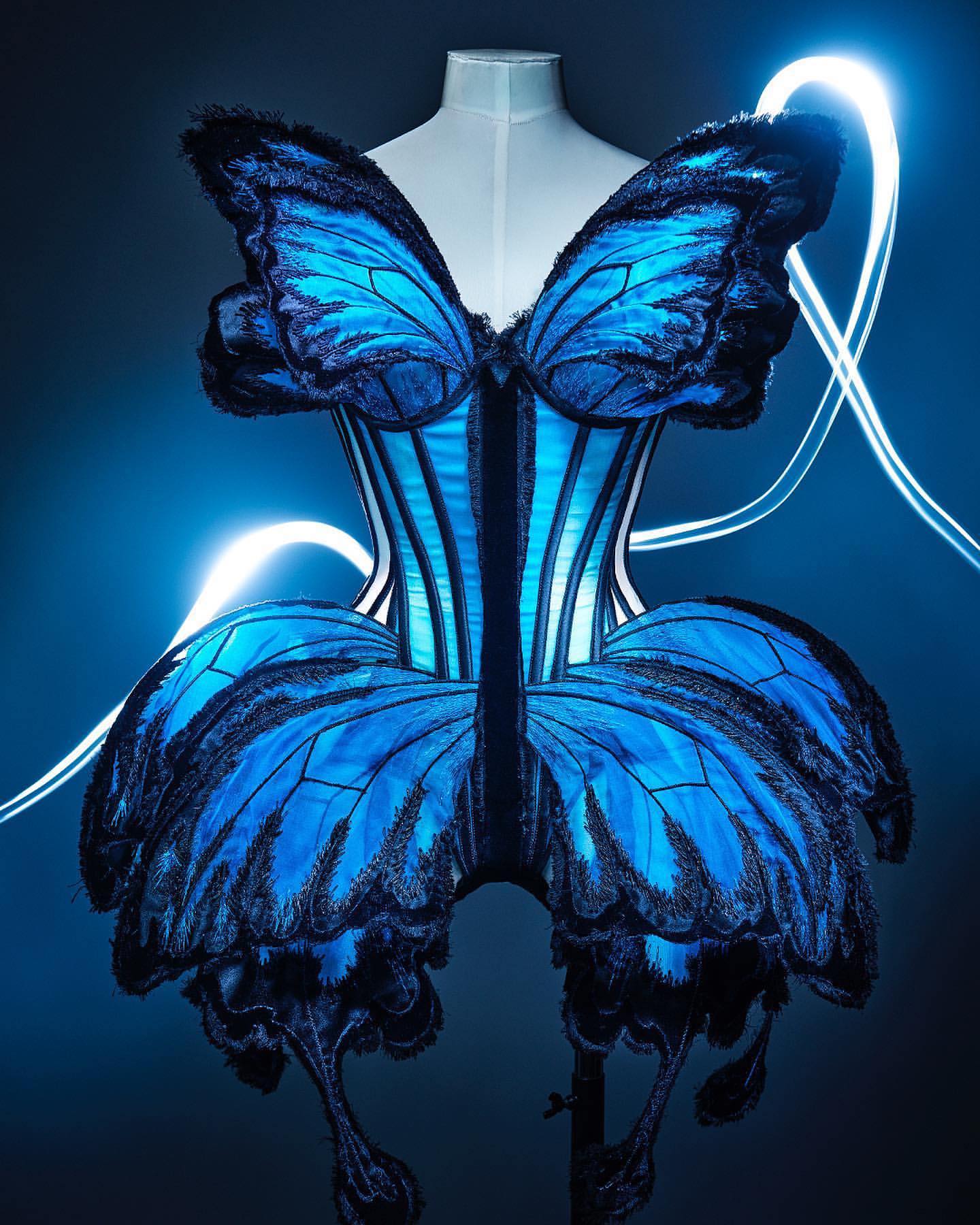 Butterfly-inspired corset dress on a mannequin, showcasing a vibrant blue and black design with delicate, wing-like detailing against a gradient blue background.