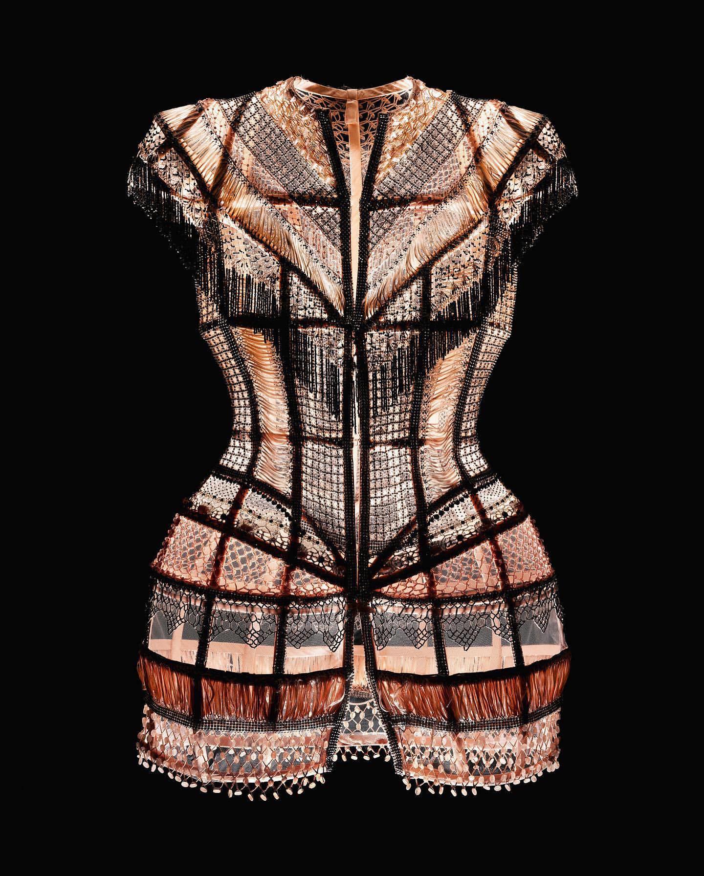 Intricate couture dress with copper and black fringe details against a black background, highlighting fashion design and craftsmanship.