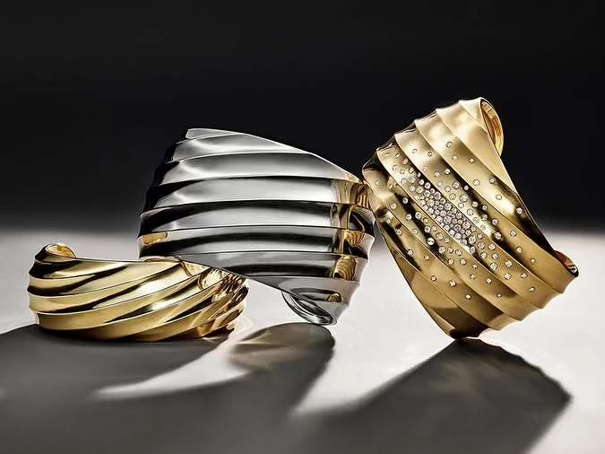 Three sculptural bangles: two gold, one silver. The gold bangle on the right features crystal embellishments. Studio lighting, dark background.