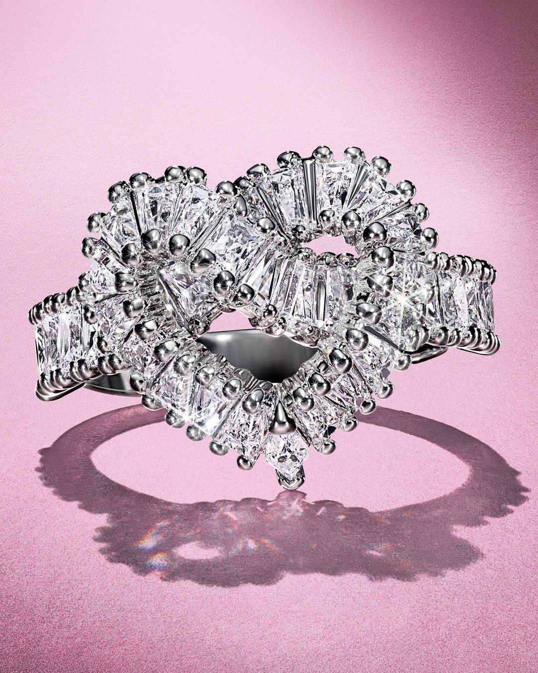 Close-up of a heart-shaped diamond ring featuring baguette-cut diamonds and bead details, set against a soft pink background.