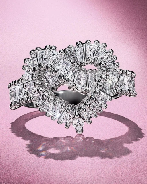 Close-up of a heart-shaped diamond ring featuring baguette-cut diamonds and bead details, set against a soft pink background.